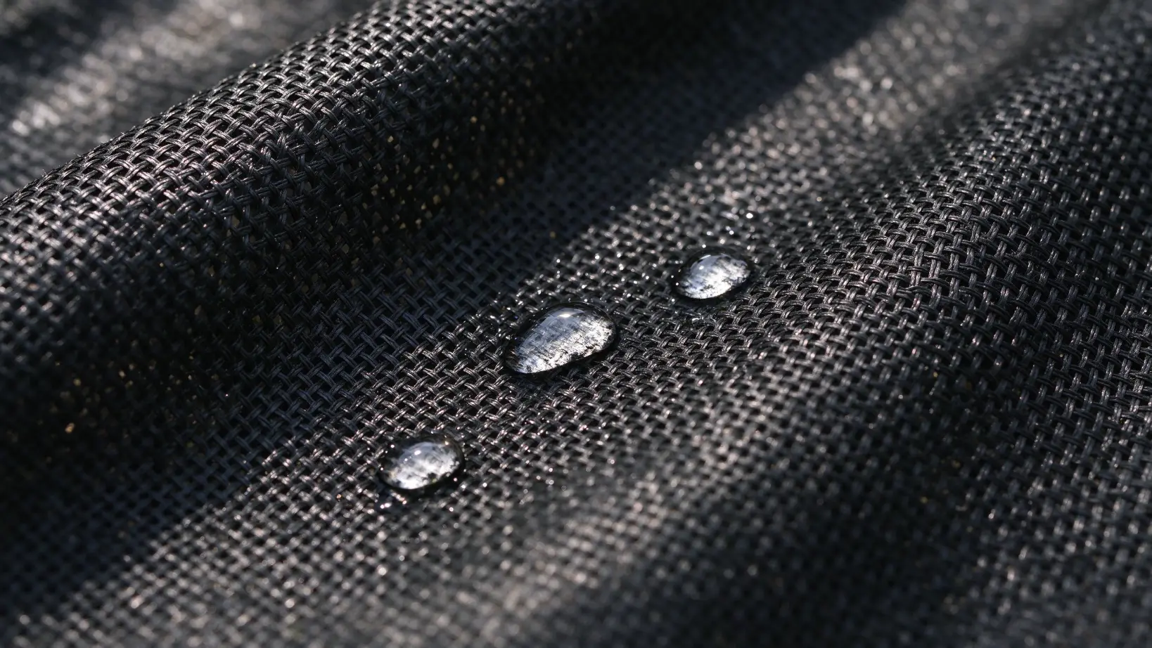 > Close-up macro of black woven polypropylene weed control fabric texture with water droplets penetrating through the grid mesh, showing permeability and UV-stabilized weave structure